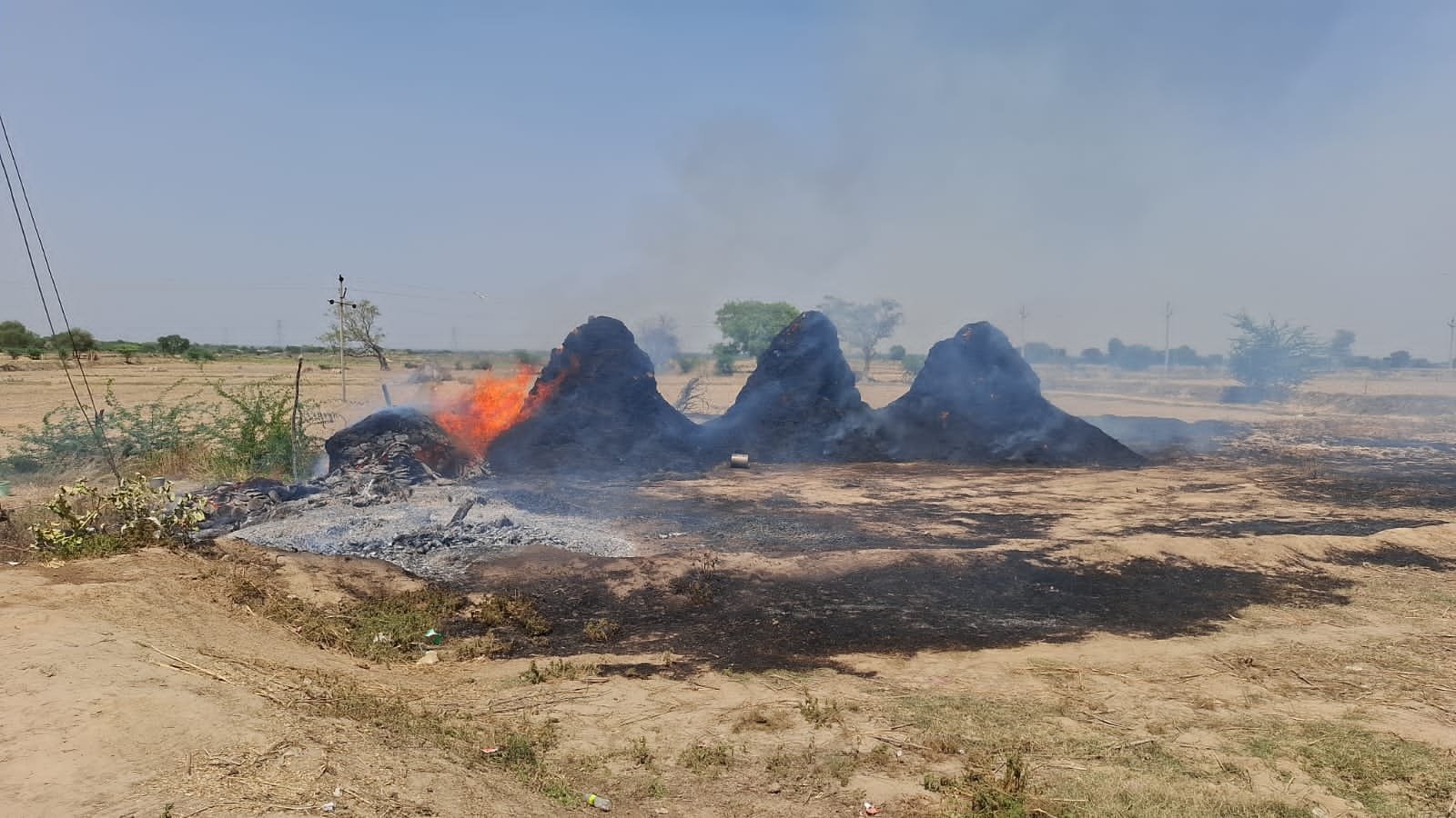 A fire incident in Babarpur village under Fatehabad area where a tube well room with a thatched roof caught fire due to a short circuit. Household items including grain, cooler, and bicycle were burnt. Villagers are seen trying to control the fire while smoke and flames spread across the structure.