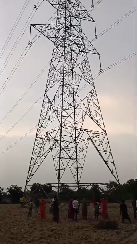 A young man sitting on a high tension electricity tower in Fatehabad while police and villagers gather below.